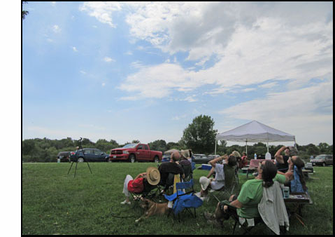 Picture shows a group of about a dozen lawn chairs, facing to our left, most of them with someone sitting and leaning back, looking up in the sky.  The sky is clear blue to our left, with the rest of the sky filled with thin white clouds.
